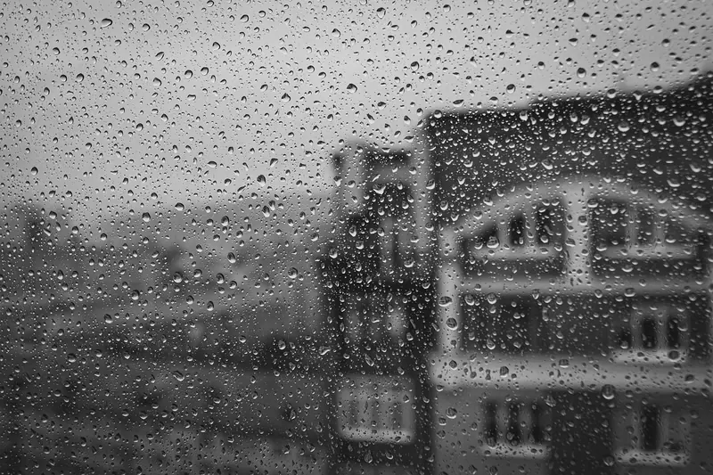 Vidro de janela coberto de gotas de chuva, com prédios desfocados ao fundo, em preto e branco