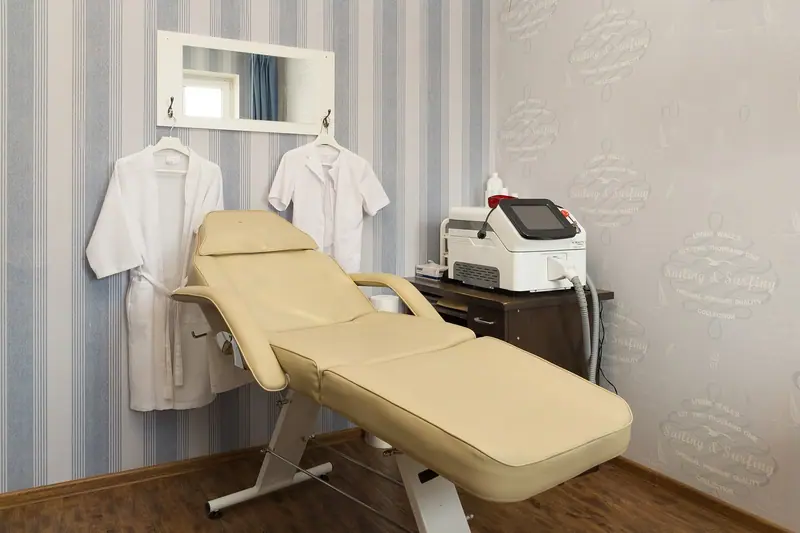 Sala de estética com cadeira reclinável bege, aparelho sobre o carrinho e jalecos pendurados