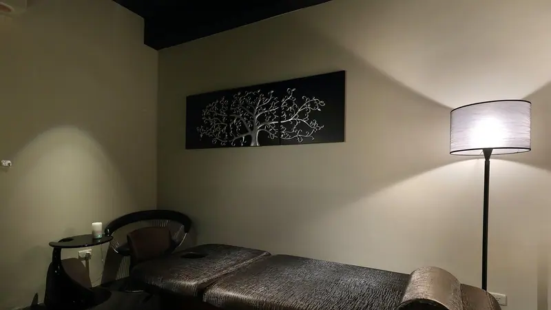 Sala de massagem à meia-luz, com maca escura, luminária de pé acesa e quadro na parede