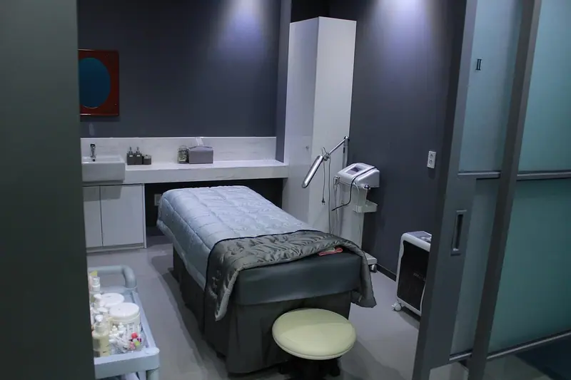 Sala de tratamento com maca, parede cinza escura e porta de vidro fosco para o corredor