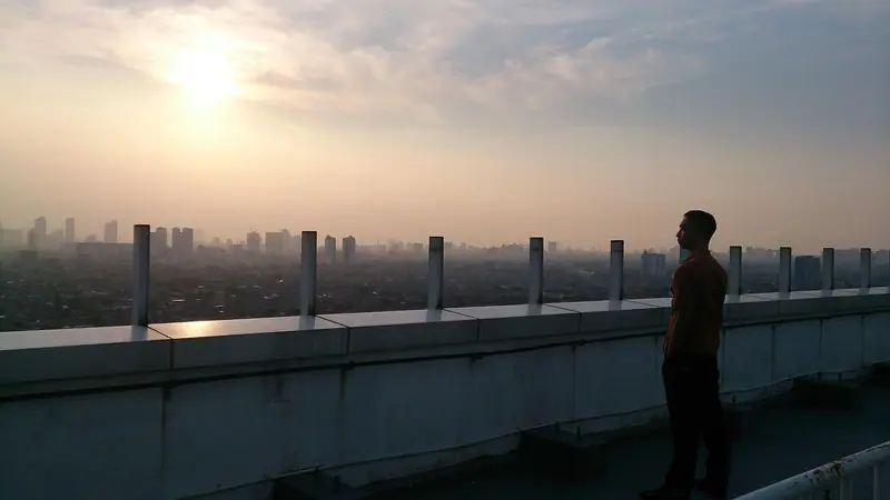 Pessoa de pé num terraço alto, de costas para a câmera, olhando a cidade sob o sol do fim da tarde