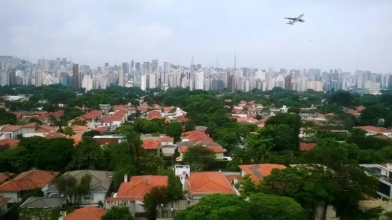 Vista do alto de São Paulo, com casas entre árvores, prédios no horizonte e um avião no céu