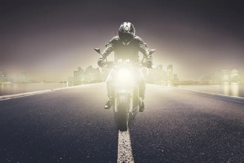 Motociclista de capacete com o farol aceso numa avenida à noite