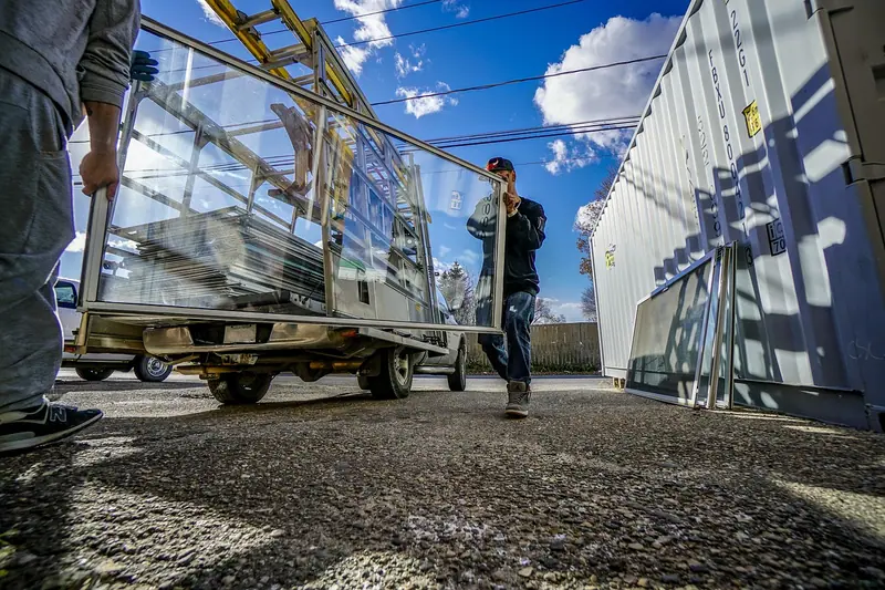 Dois trabalhadores de longe empurrando um carrinho com painéis de vidro ao lado de um galpão