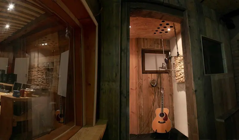 Cabine de gravação revestida de madeira, com violão e microfone, vista da sala de controle