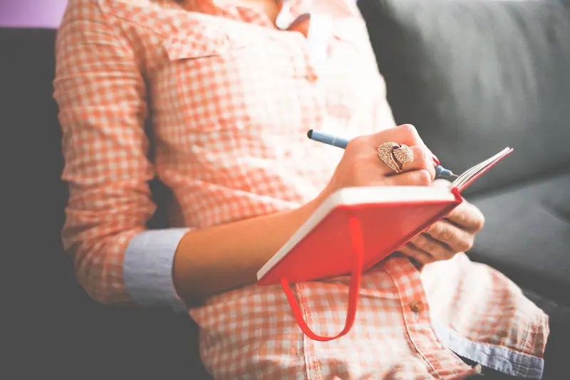 Pessoa sentada no sofá escrevendo num caderno vermelho, sem o rosto aparecendo