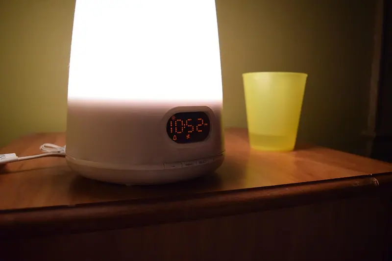 Luminária acesa sobre o criado-mudo, ao lado de um copo amarelo, num quarto à meia-luz