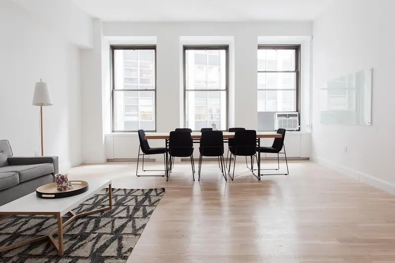 Sala clara e quase vazia, com mesa de jantar, sofá e três janelas na parede do fundo
