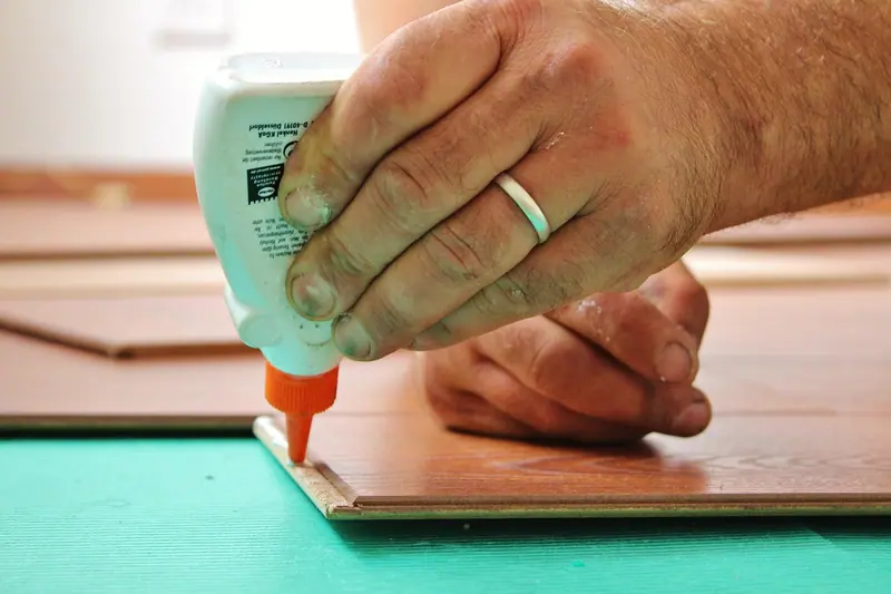 Mão aplicando cola na borda de uma régua de piso laminado durante a instalação