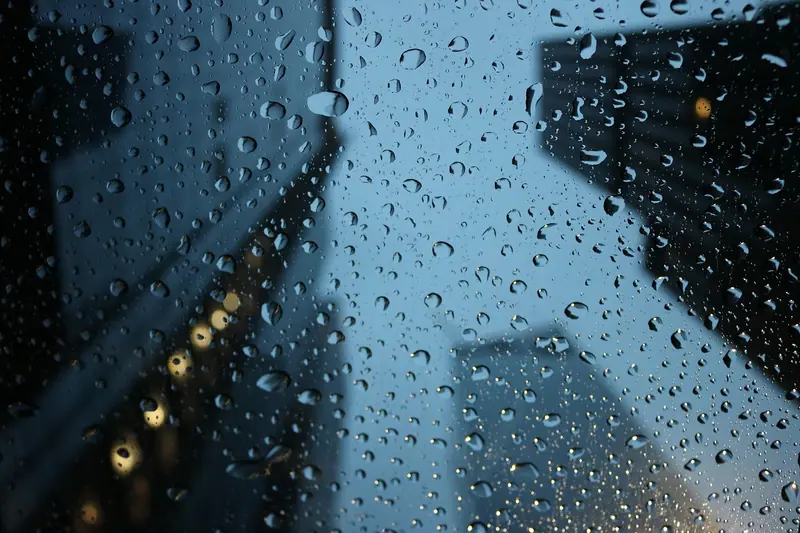 Gotas de chuva no vidro de uma janela, com prédios altos desfocados ao fundo