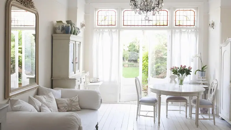 Sala branca e elegante com sofá, lustre de cristal, mesa redonda e portas de vidro abertas para o jardim