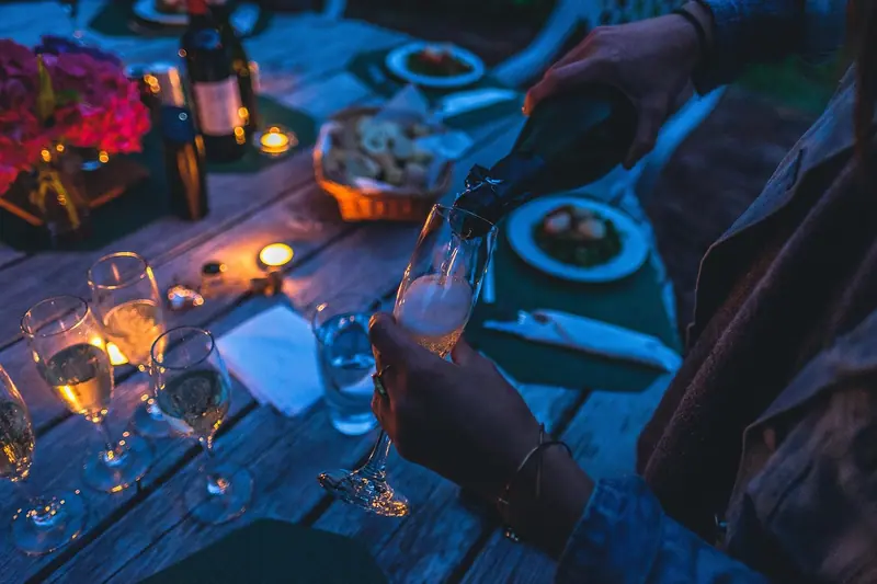Mãos servindo vinho em taças numa mesa de madeira iluminada por velas à noite