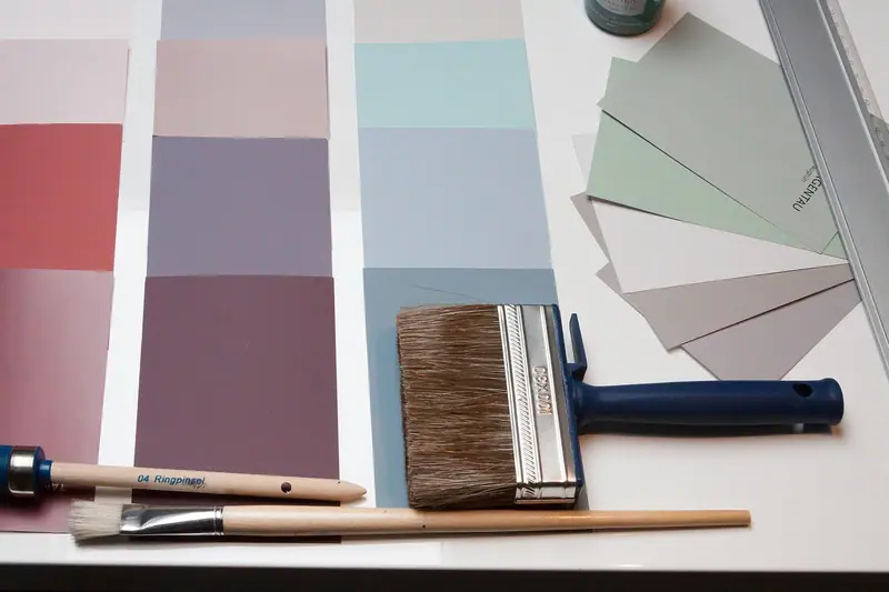 Cartelas de cores em tons de rosa e verde, com um pincel largo e um pincel fino sobre a mesa