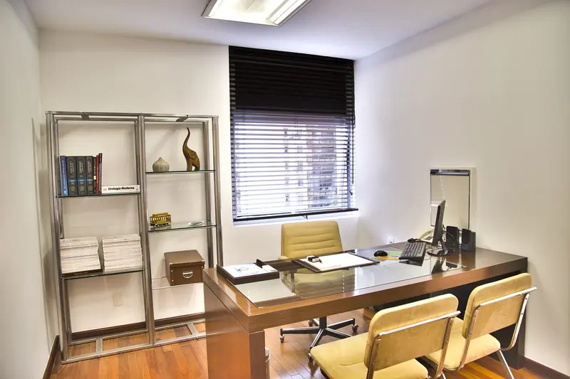 Sala de atendimento com mesa de madeira, cadeiras, estante e janela com persiana