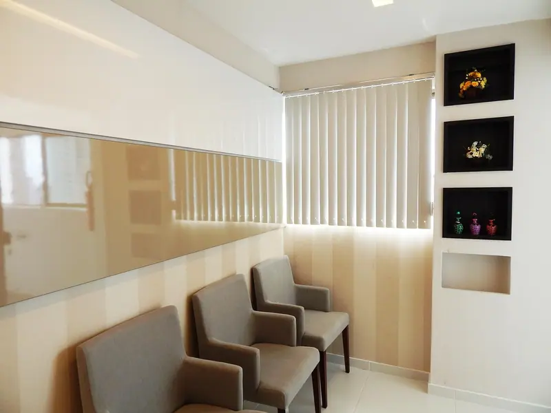 Sala de espera clara com poltronas cinza enfileiradas junto a uma janela com persiana