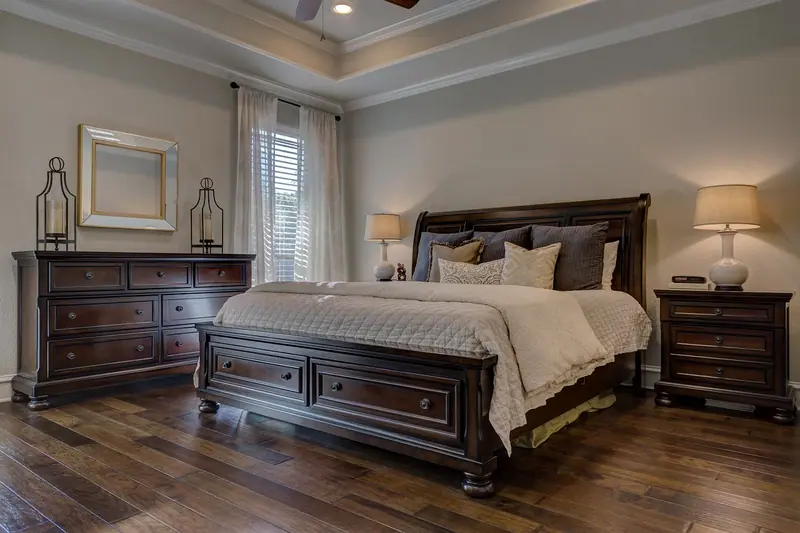 Quarto com cama de madeira escura, abajures acesos e cortinas claras nas janelas