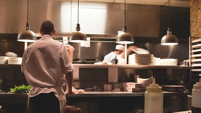 Cozinha profissional de inox em plena atividade, com cozinheiros de costas e luminárias pendentes acesas