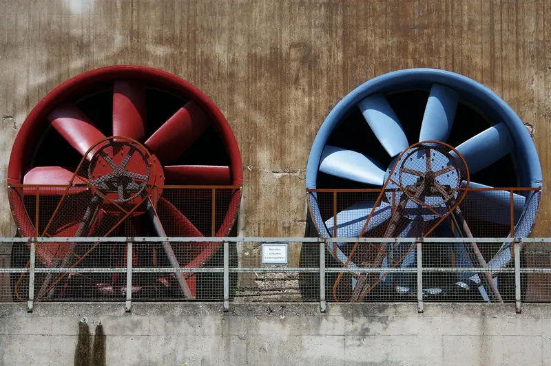 Dois grandes ventiladores industriais, um vermelho e outro azul, presos numa parede de concreto