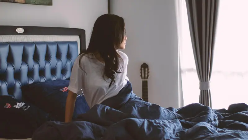 Mulher sentada na cama, de lado, olhando para a janela com cortinas cinza num quarto claro
