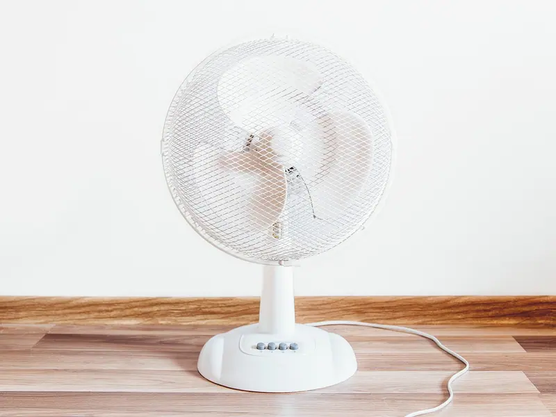 Ventilador branco de mesa sobre o piso de madeira, encostado numa parede clara
