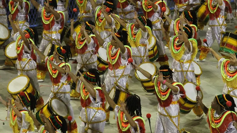 Bateria de escola de samba desfilando, com dezenas de ritmistas fantasiados tocando tambores
