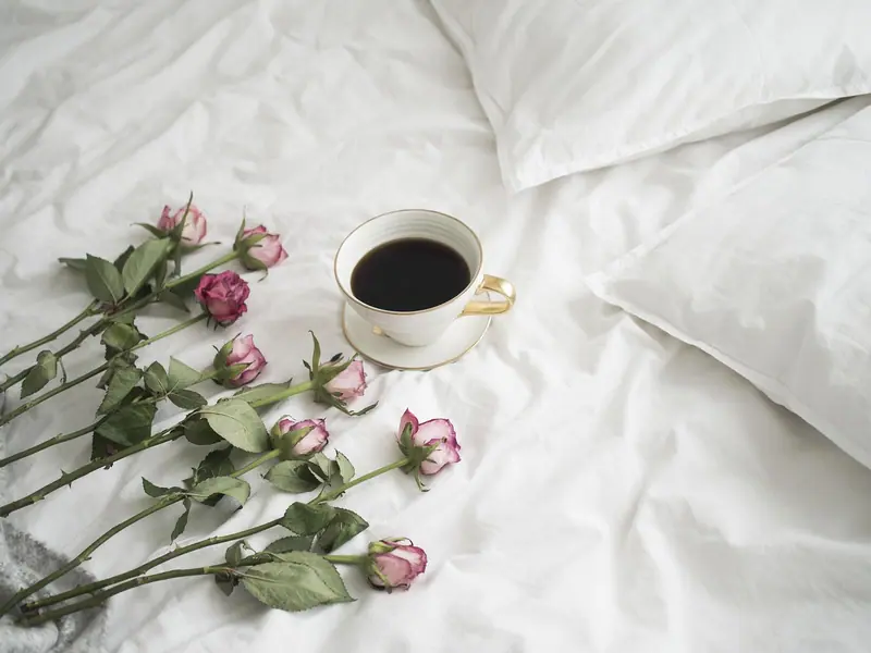 Xícara de café sobre lençóis brancos, ao lado de rosas cor-de-rosa, numa cama arrumada