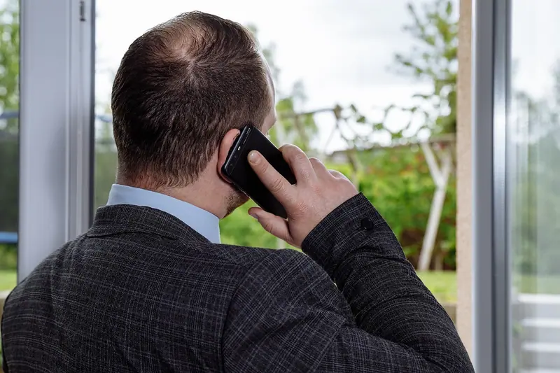 Homem de costas falando ao celular perto de uma janela de vidro com vista para árvores