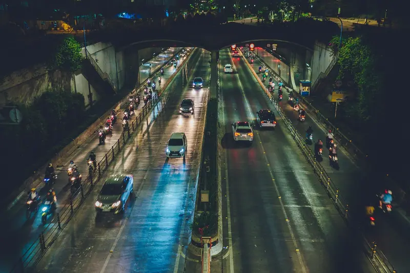 Avenida molhada à noite, vista do alto, com carros e motos passando entre árvores
