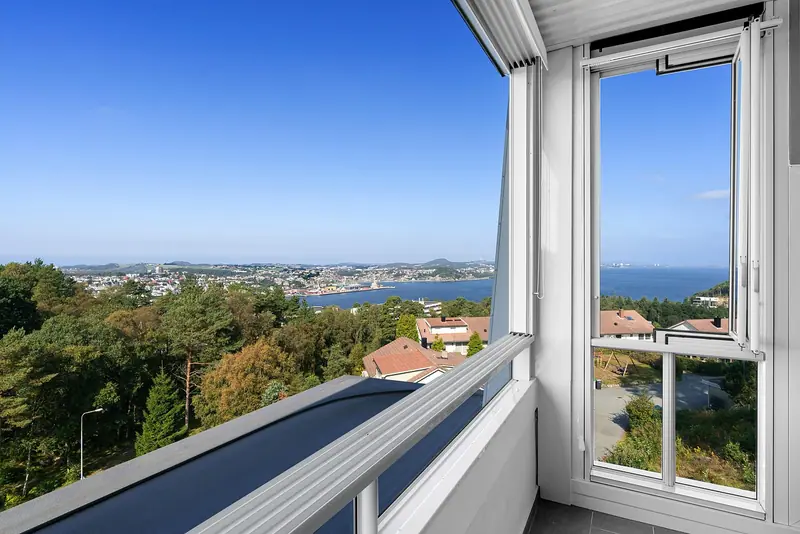 Varanda com piso escuro, gradil branco e uma janela alta de vidro, com vista para árvores e casas