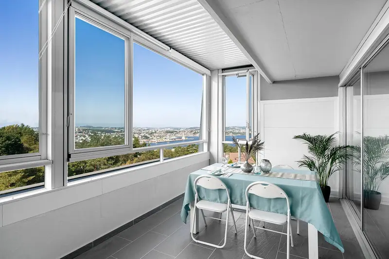 Varanda envidraçada com mesa posta, cadeiras dobráveis e plantas, com vista para a cidade