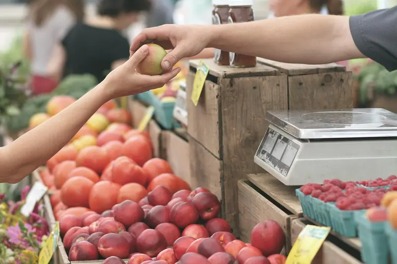 Mãos de cliente e de feirante trocando uma fruta sobre caixotes, com uma balança ao lado