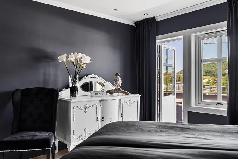 Quarto de parede escura com cama, cômoda branca e uma janela de vidro clara ao fundo