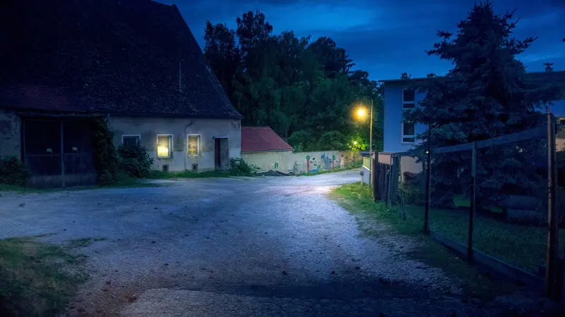 Rua de terra batida à noite, iluminada por um poste aceso, com uma casa de janelas acesas ao lado