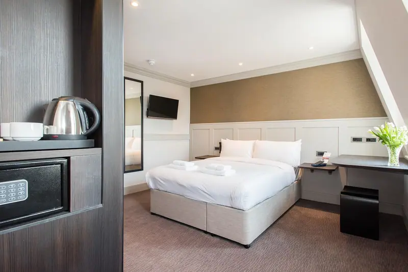 Entrada de quarto de hotel com chaleira e cofre no armário e cama de casal ao fundo
