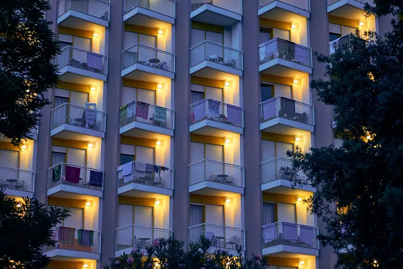 Fachada de prédio com fileiras de varandas iluminadas no começo da noite, entre árvores