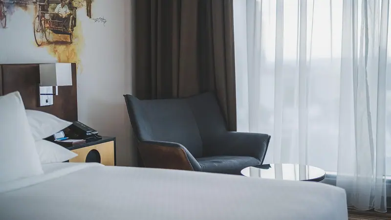 Quarto de hotel com cama de lençóis brancos, poltrona cinza e cortina clara filtrando a luz