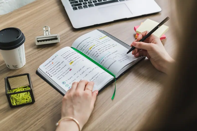 Mãos escrevendo anotações num caderno sobre a mesa, ao lado de um notebook