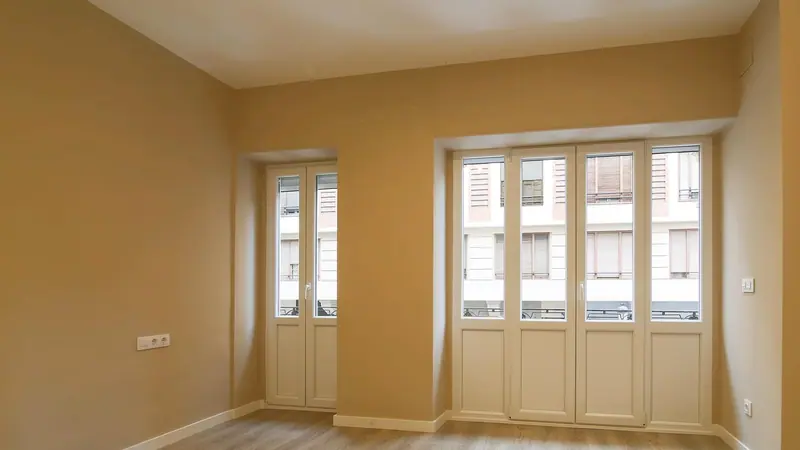 Sala vazia de paredes amarelas, com porta e janelas de vidro voltadas para os prédios da rua