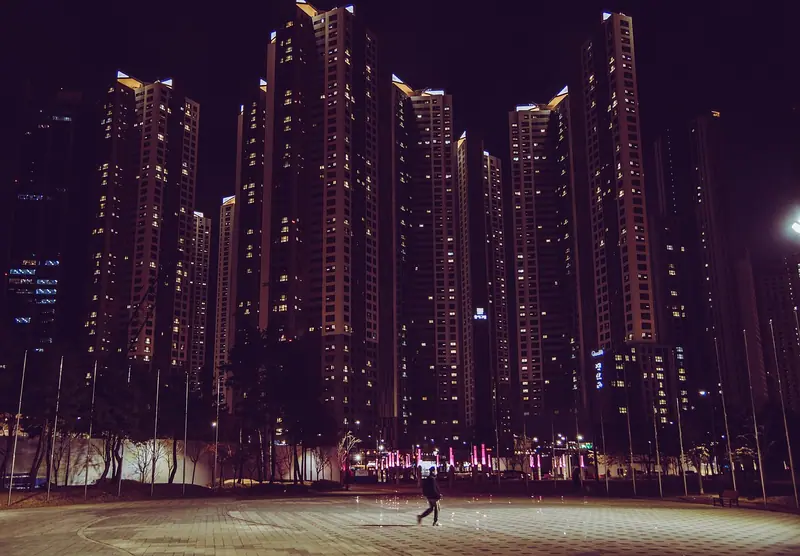 Torres residenciais com janelas acesas à noite e uma pessoa pequena caminhando na praça em frente