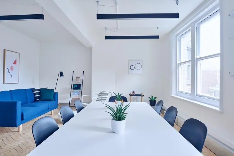 Sala ampla e clara com sofá azul, mesa branca comprida e janelas grandes