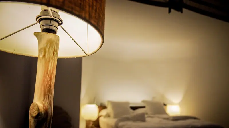 Abajur de madeira aceso em primeiro plano e uma cama ao fundo, num quarto à meia-luz