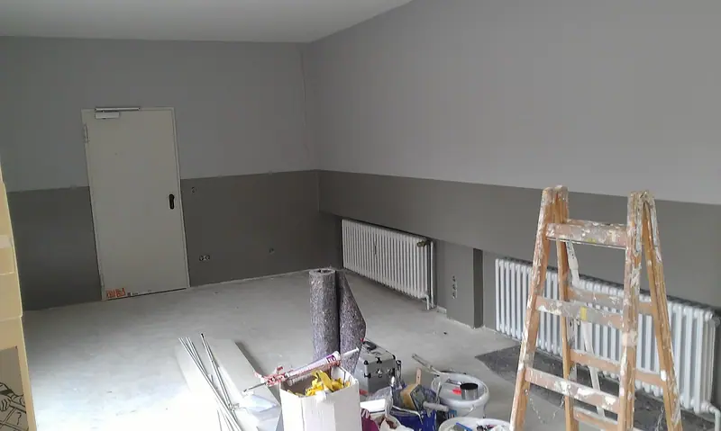 Cômodo em obra com paredes cinza, uma porta ao fundo, escada de pintor e materiais no chão