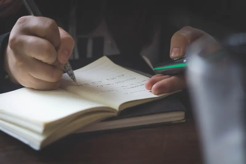 Mãos anotando com caneta num caderno aberto sobre a mesa, em luz baixa