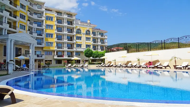 Piscina azul com espreguiçadeiras ao pé de um prédio de vários andares com varandas, num dia de sol