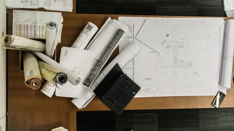 Plantas de projeto enroladas e abertas sobre uma mesa de madeira, ao lado de um notebook