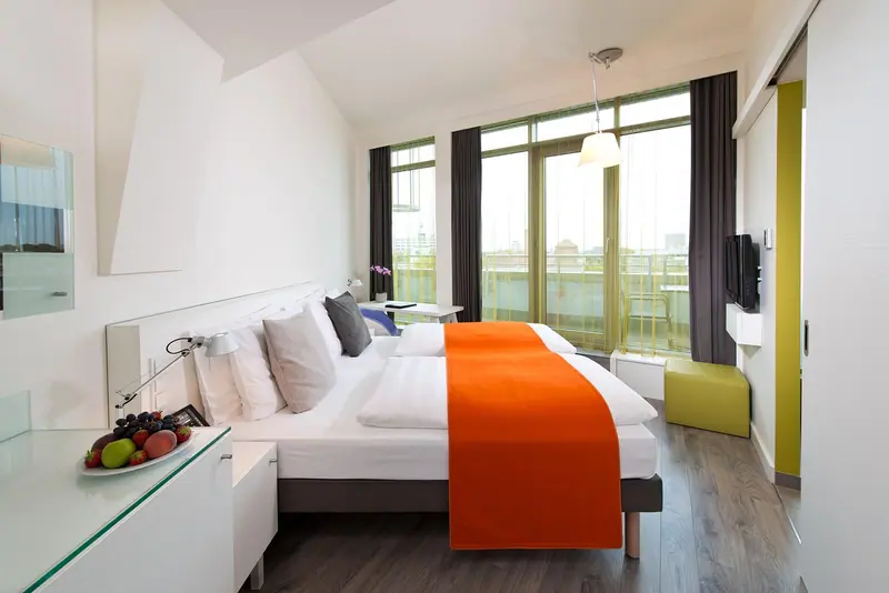 Quarto de hotel claro com cama arrumada, manta laranja e janelas grandes com cortinas