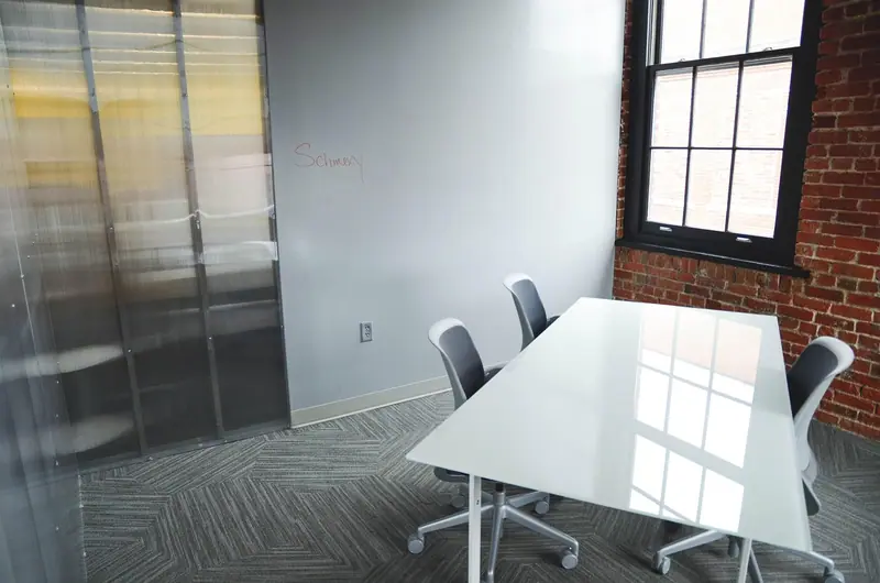 Sala de reunião pequena com mesa branca e cadeiras, separada do corredor por divisória de vidro