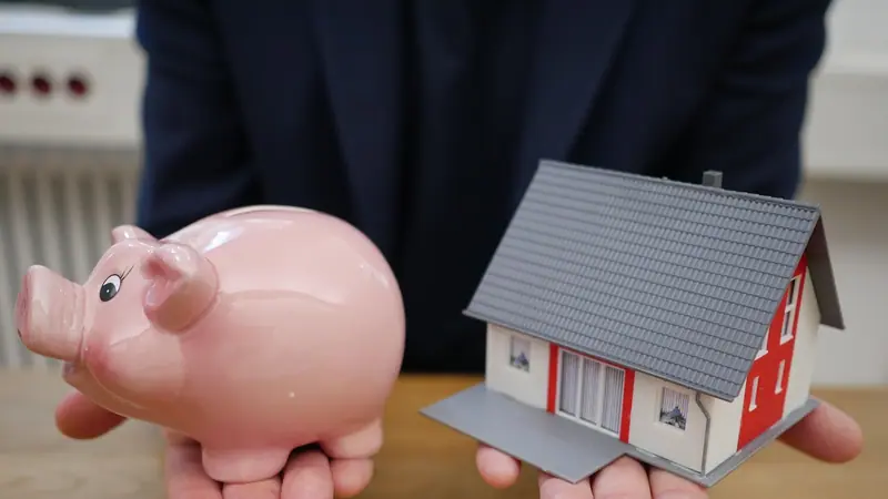 Mãos estendidas segurando um cofrinho de porquinho rosa e uma pequena maquete de casa