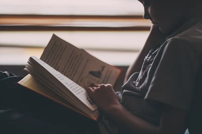 Pessoa lendo um livro ao lado de uma persiana, em contraluz