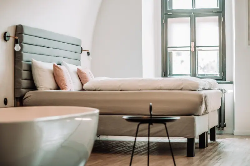 Quarto com cama de cabeceira estofada e uma janela alta deixando a luz do dia iluminar o ambiente